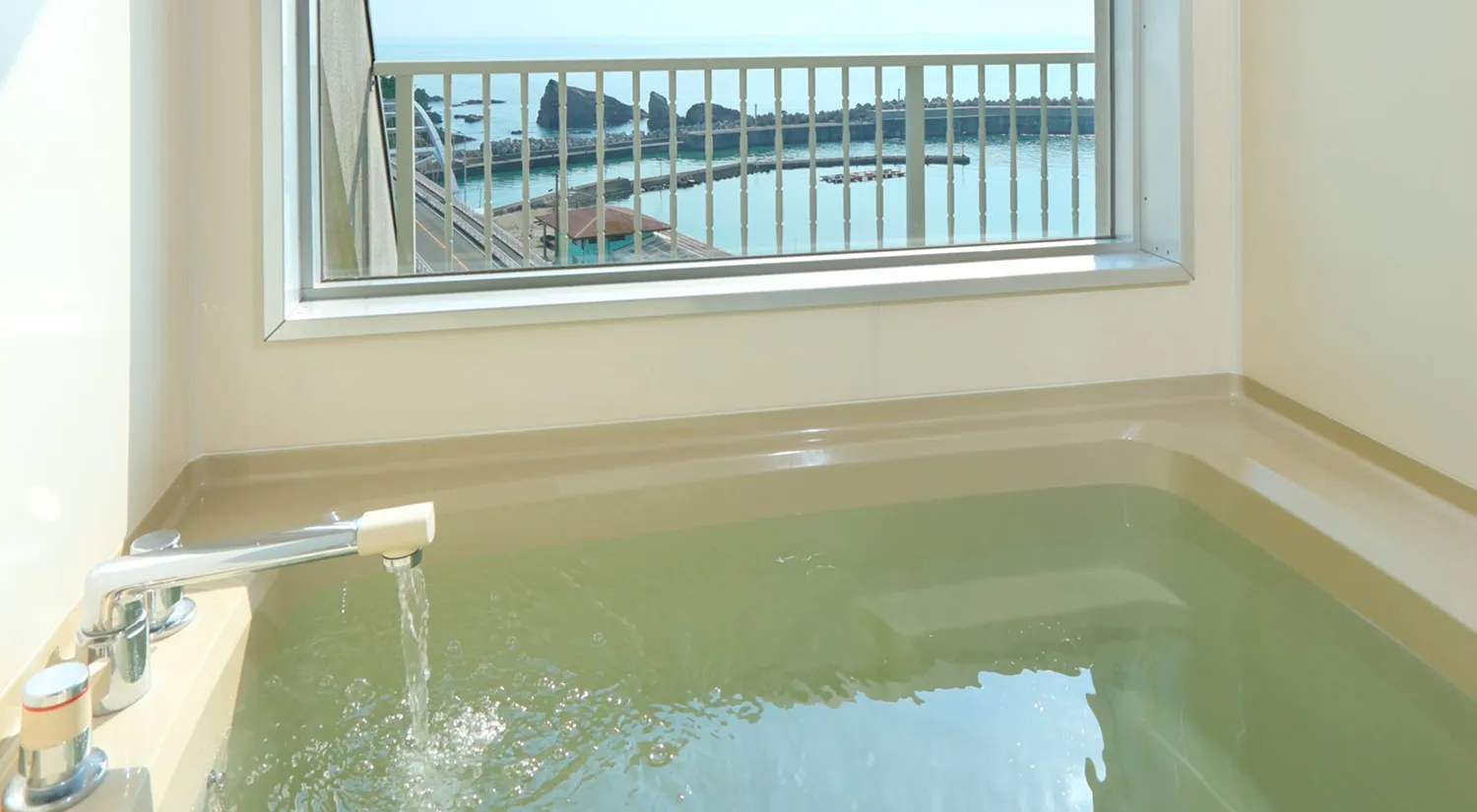 窓から海と漁港が見える浴室で、浴槽にお湯が注がれています。
